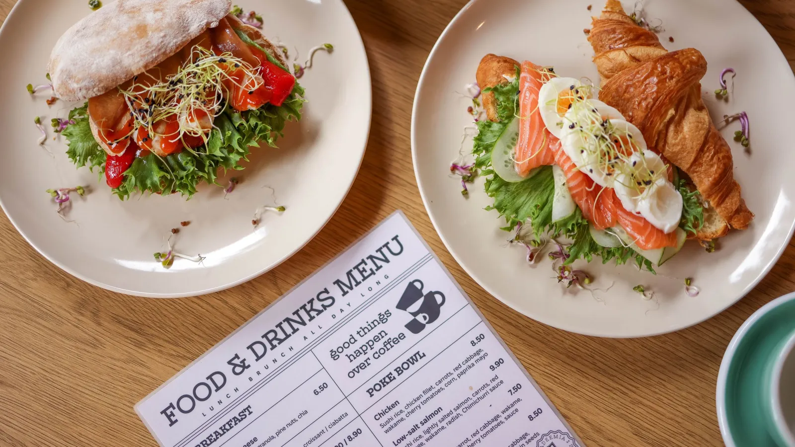 Printed food and drinks menu on a wooden table next to two plates with open sandwiches