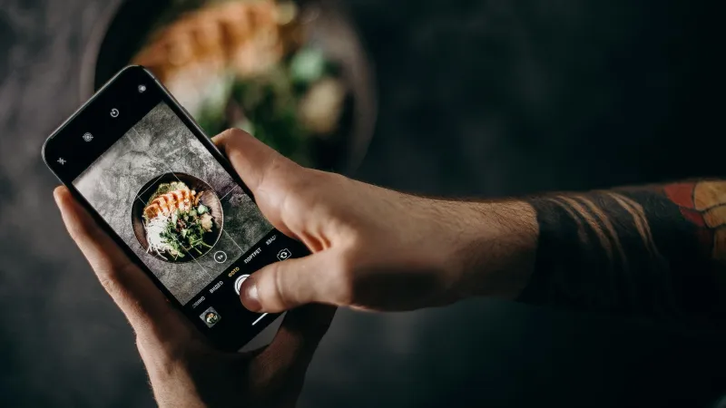 Hand holding a smartphone and photographing a plated dish on a dark table