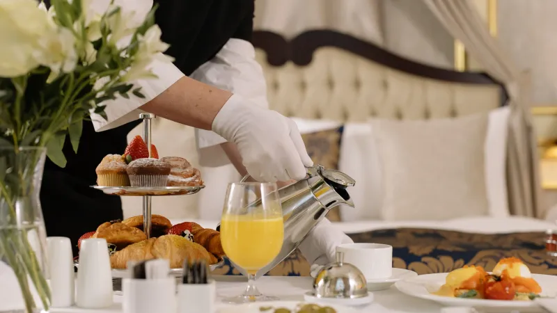 Waiter in white gloves pouring coffee beside a room service breakfast with pastries and orange juice
