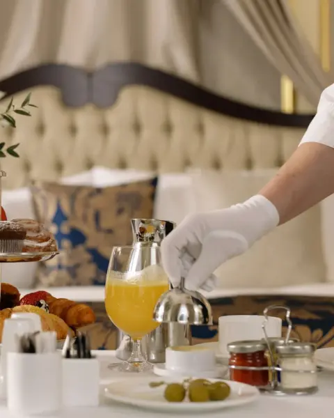 A waiter in white gloves serves breakfast with orange juice and pastries on a hotel bed